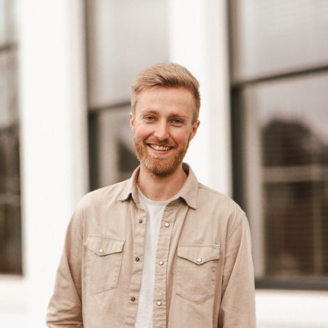 Man in a color-block sweater leaning against a white wall with arms crossed, smiling.