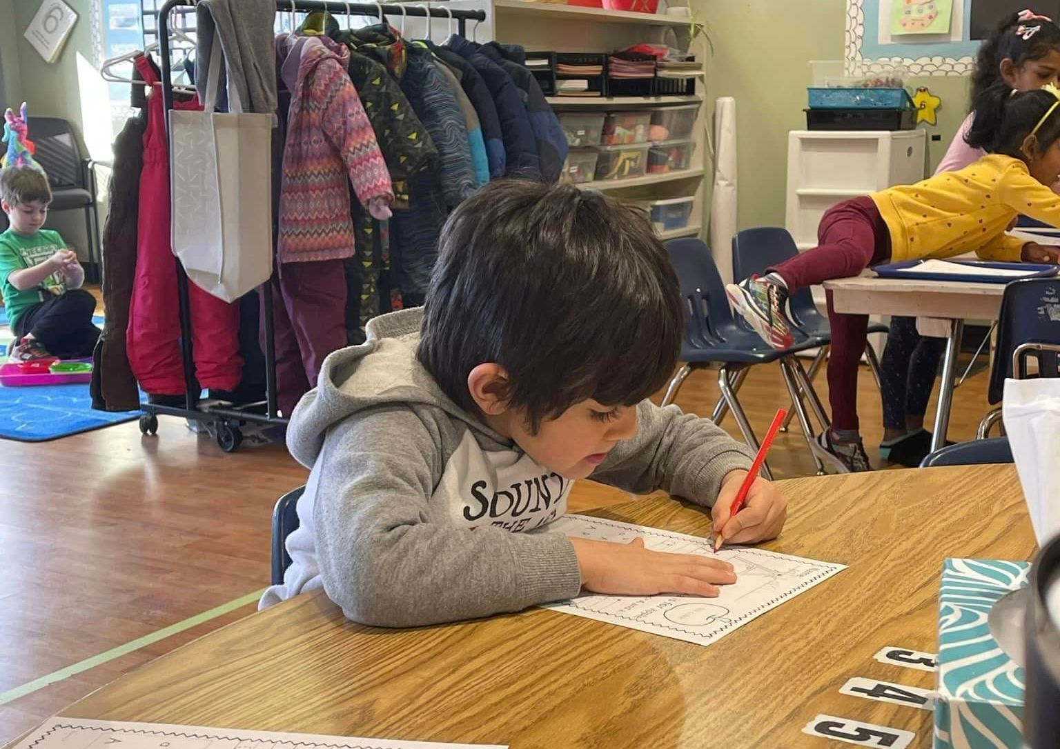 A young boy is sitting at a desk in a classroom writing on a piece of paper. | Brookline, NH | Scribbles Early Learning