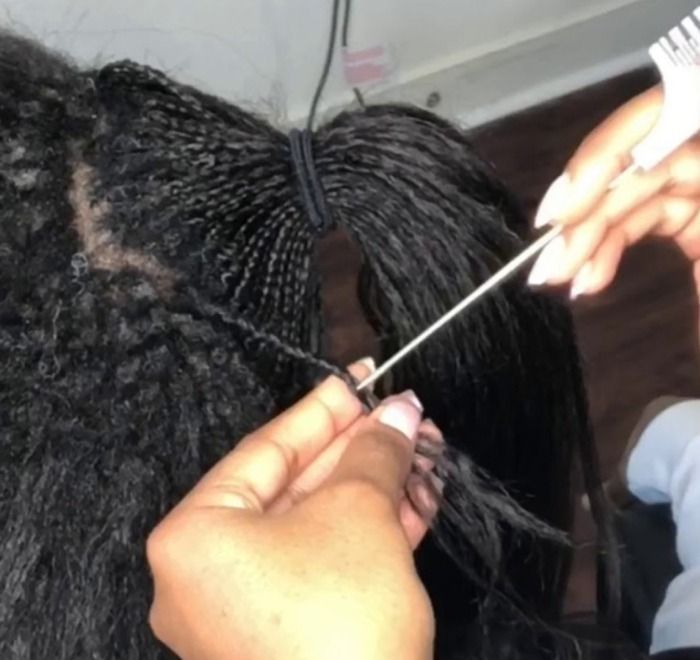 Hands braiding hair with a styling tool. Dark hair is tied back, revealing sections of tiny braids.