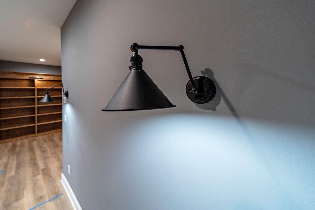 Black adjustable wall sconce on a gray wall, illuminating the surface. Wooden shelves are in the background.