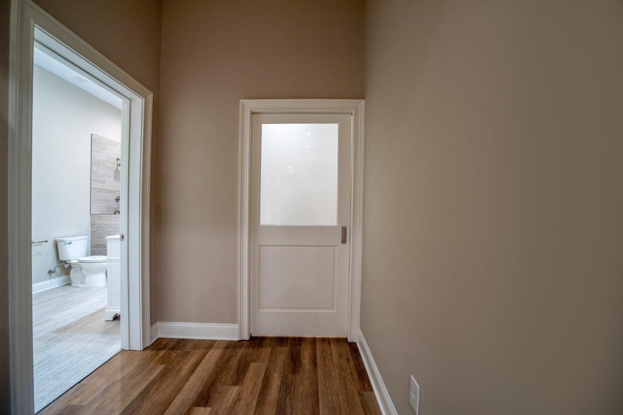 Hallway with wooden floor, two doorways, one to a bathroom, the other with a frosted glass door. Beige walls.