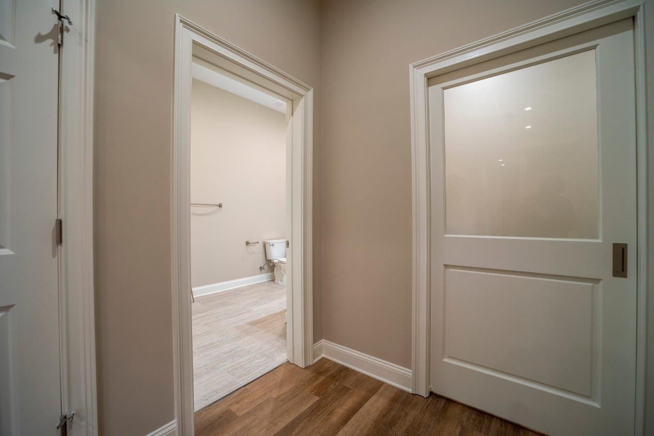 Hallway with three doors: one open to a bathroom, one closed with frosted glass, one closed. Light brown walls, wood floor.