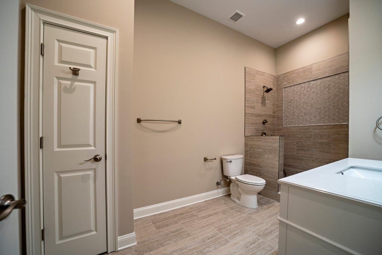 Bathroom with beige walls, white door, toilet, shower, and vanity.