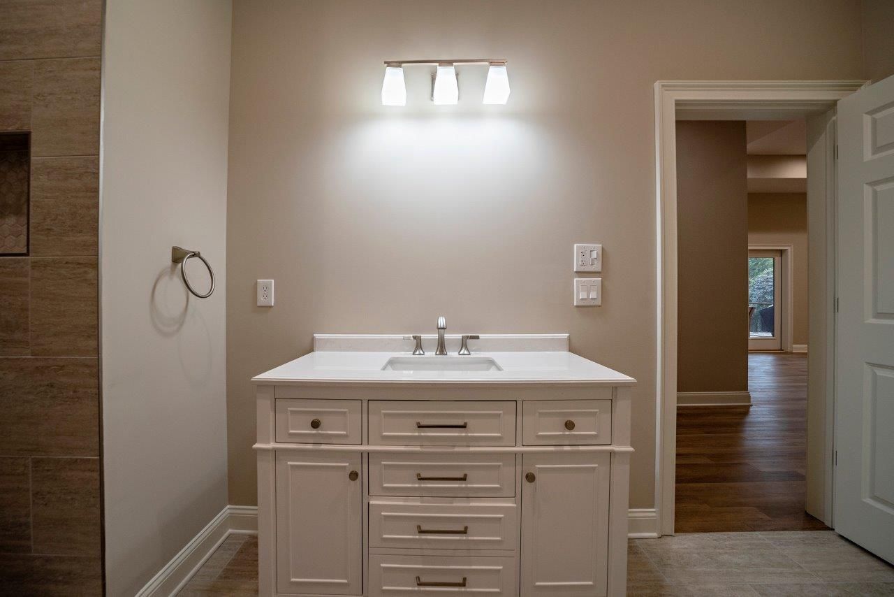 Bathroom with white vanity, sink, three-light fixture, towel ring, open doorway, and a tiled shower on the left.