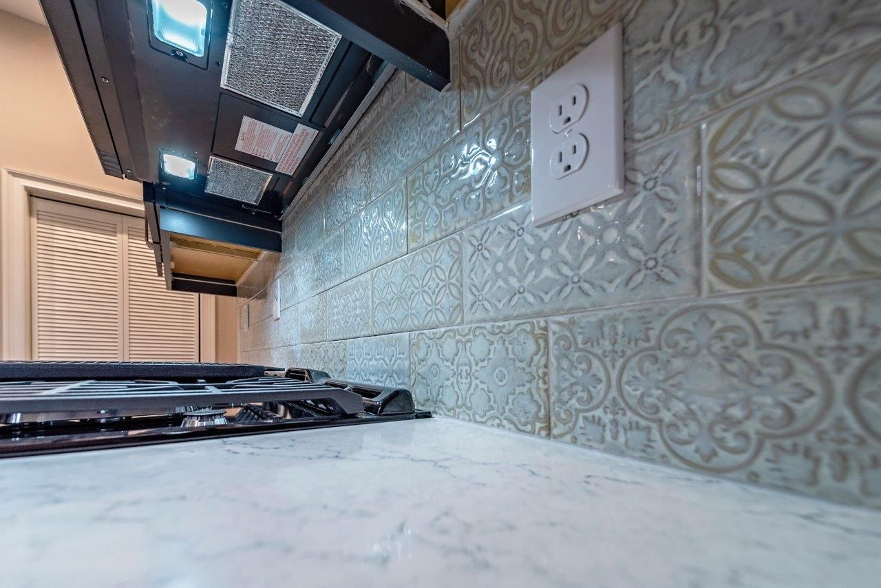 Kitchen backsplash with decorative tile, outlet, and range hood. Gray-blue tiles with white countertop.