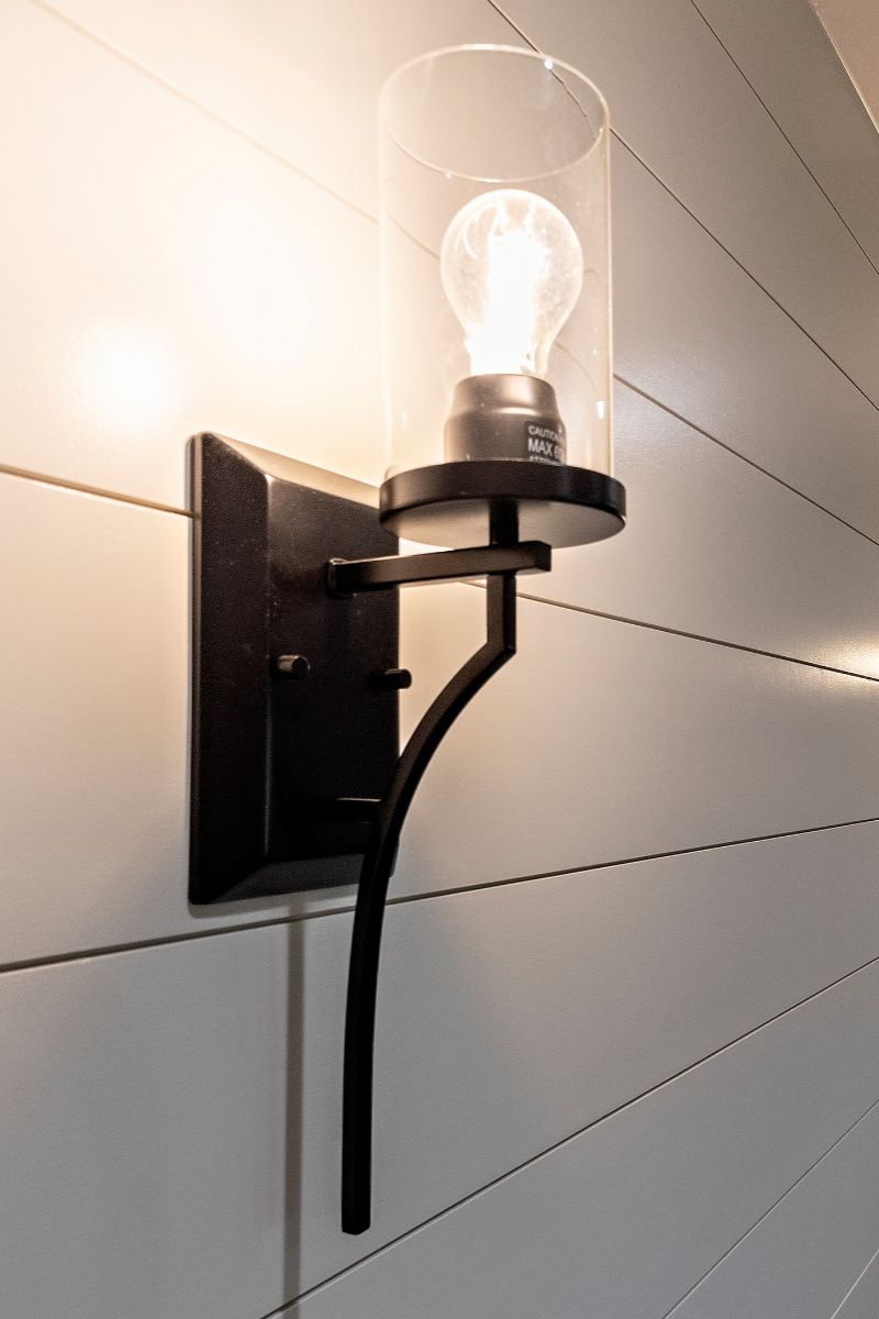 Black sconce with cylindrical glass shade and lit bulb on a white paneled wall.