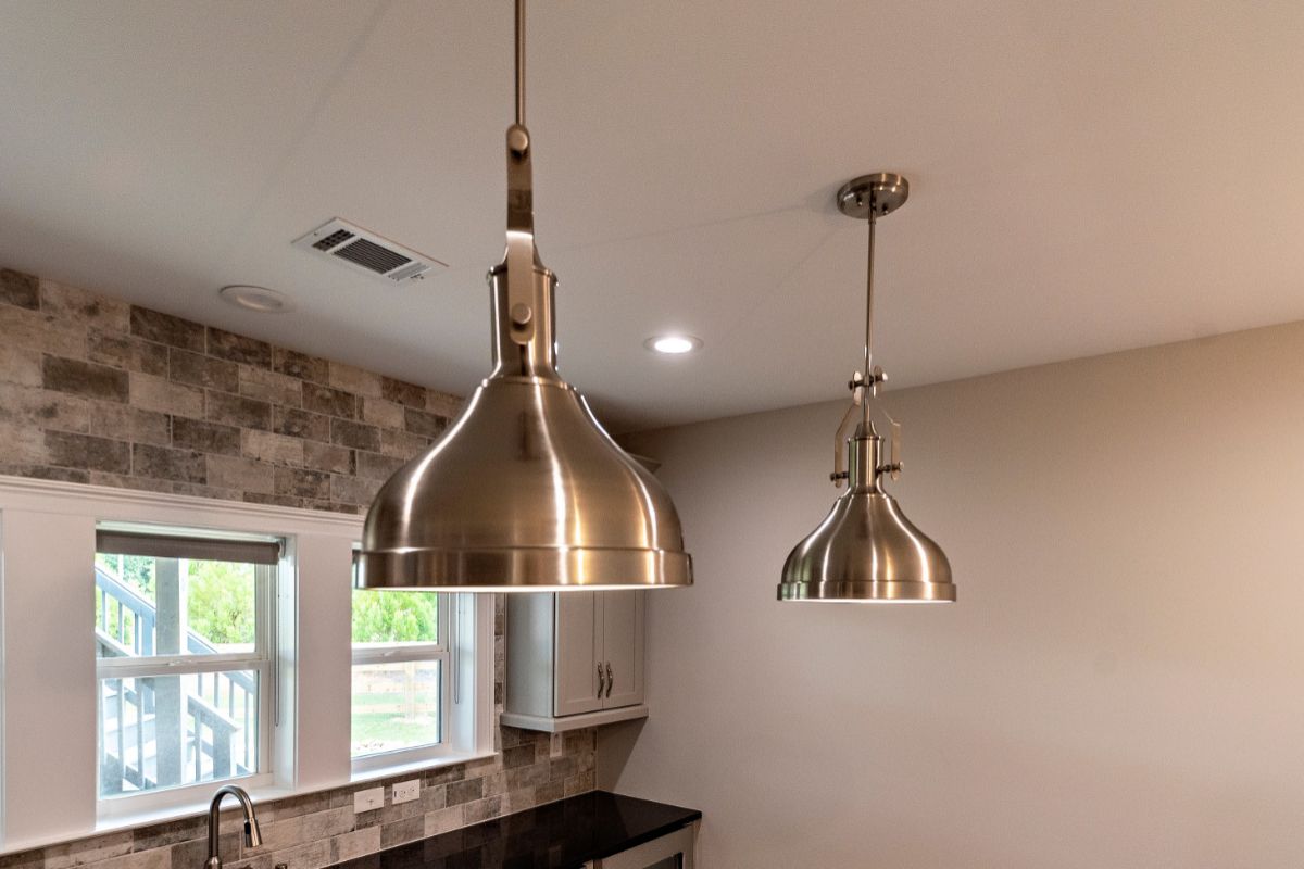 Two brushed metal pendant lights hanging from a white ceiling, above a kitchen counter with brick-like backsplash.
