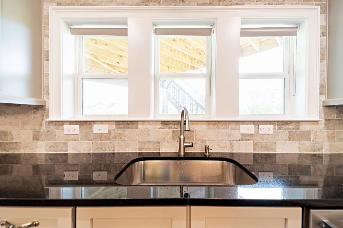 Stainless steel kitchen sink under three windows with roller shades, above a dark countertop.