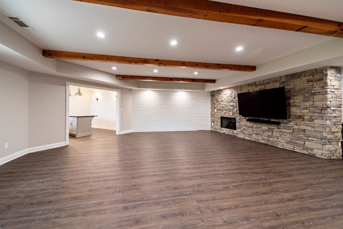 Spacious finished basement with wooden beams, stone accent wall, fireplace, TV, and wood-look flooring.