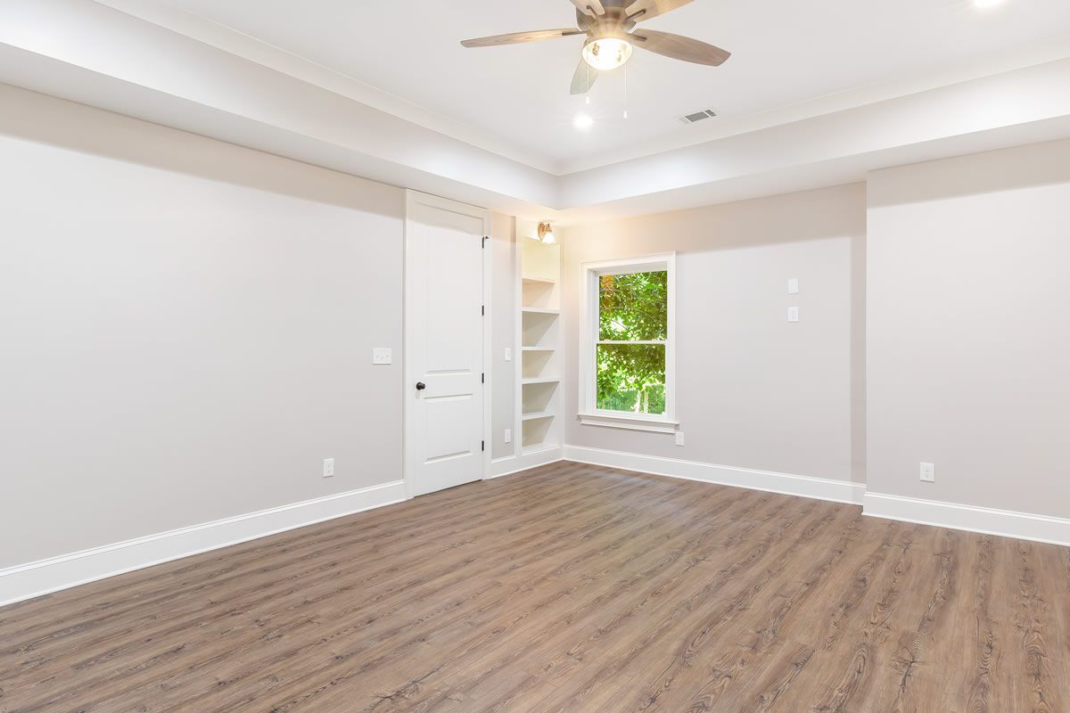 Empty bedroom with light grey walls, white trim, wood-look floor, window, built-in shelves, and ceiling fan.