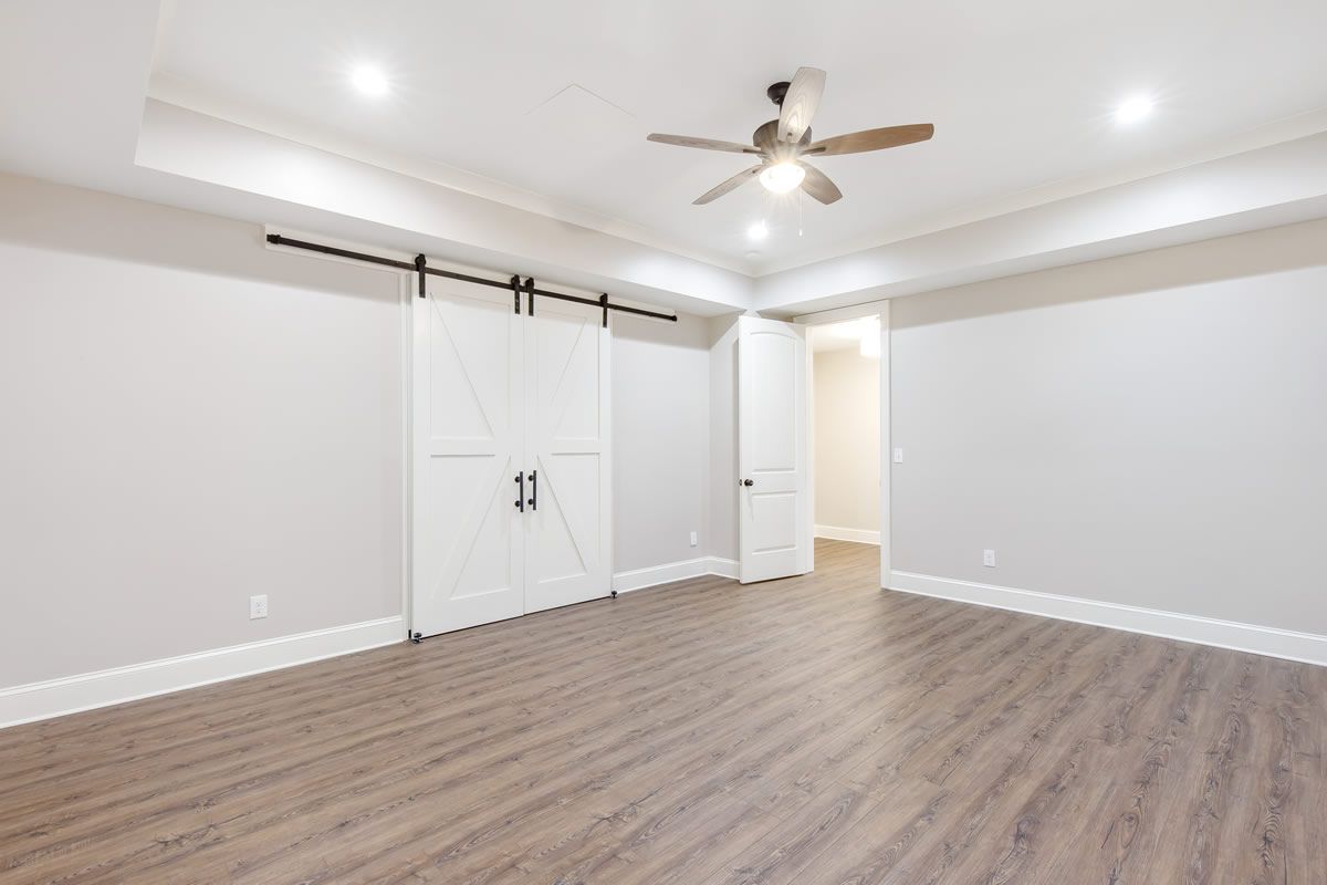 Empty, light-filled room with wood-look floor, barn doors, and recessed lighting. A ceiling fan hangs in the center.