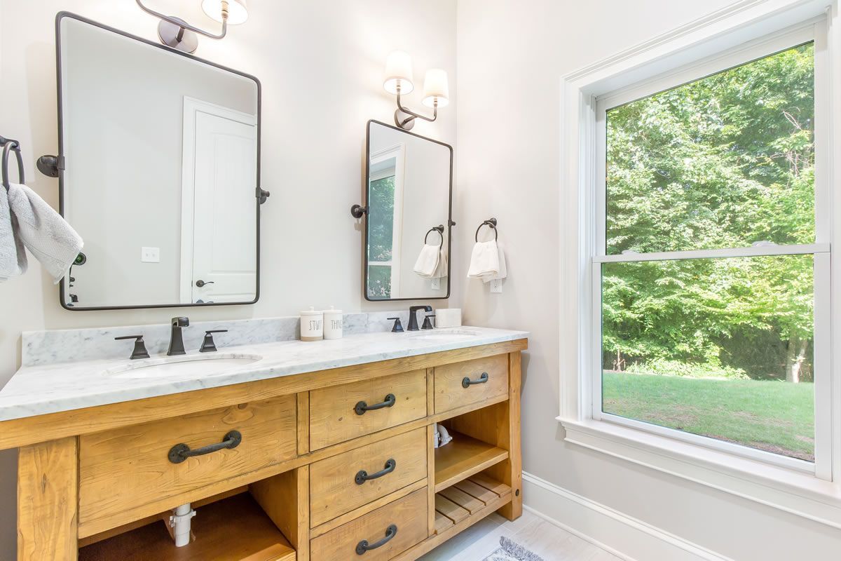 Bathroom with double vanity, two mirrors, and a window overlooking greenery. Light wood cabinet with black hardware.