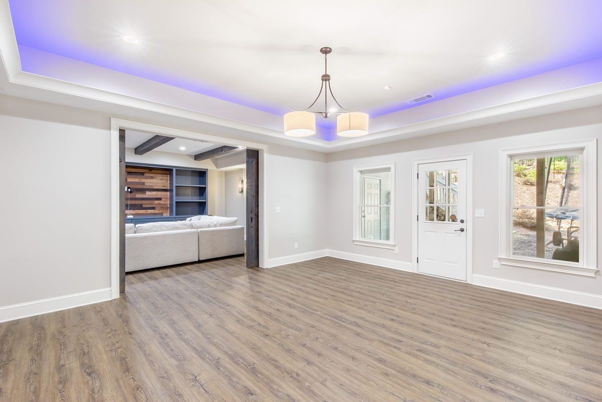 Empty, modern room with hardwood floors and bright lighting, leading to a living area with built-in shelves and a sofa.