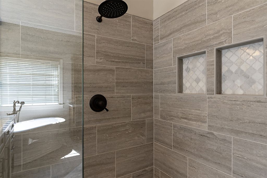 Bathroom shower with gray tiled walls, glass door, and dark fixtures. Two inset shelves with mosaic tiles.