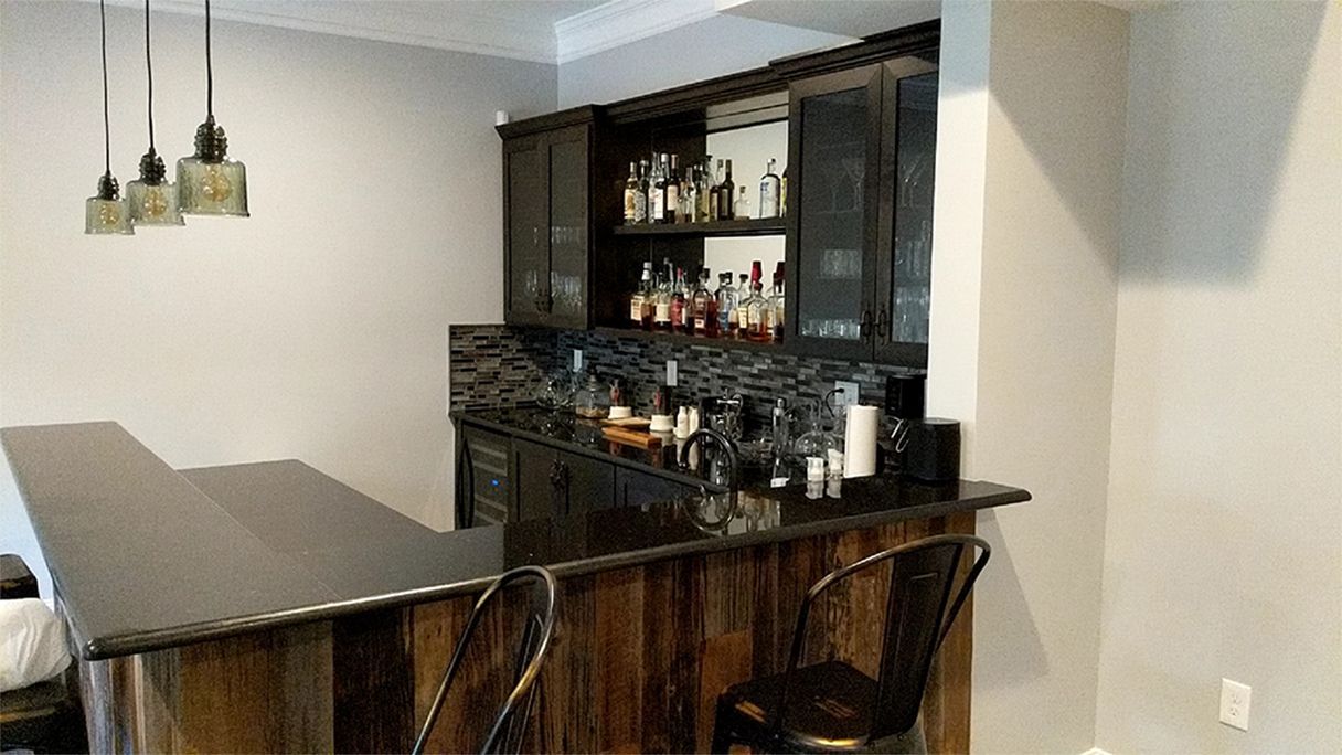 A home bar with black countertop and wood paneling. Bottles, glasses, and stools are visible.