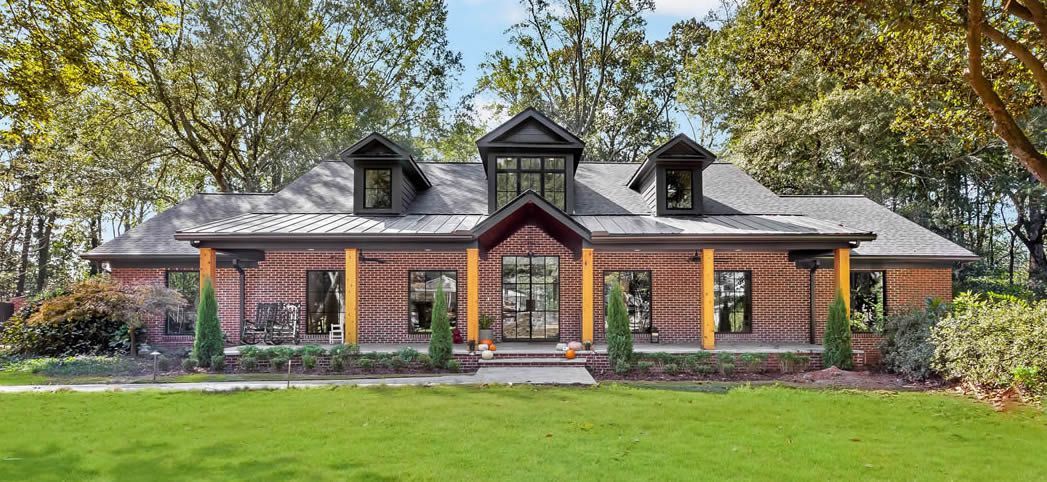 Brick house with a covered porch and dormers, set on a green lawn with trees in the background.
