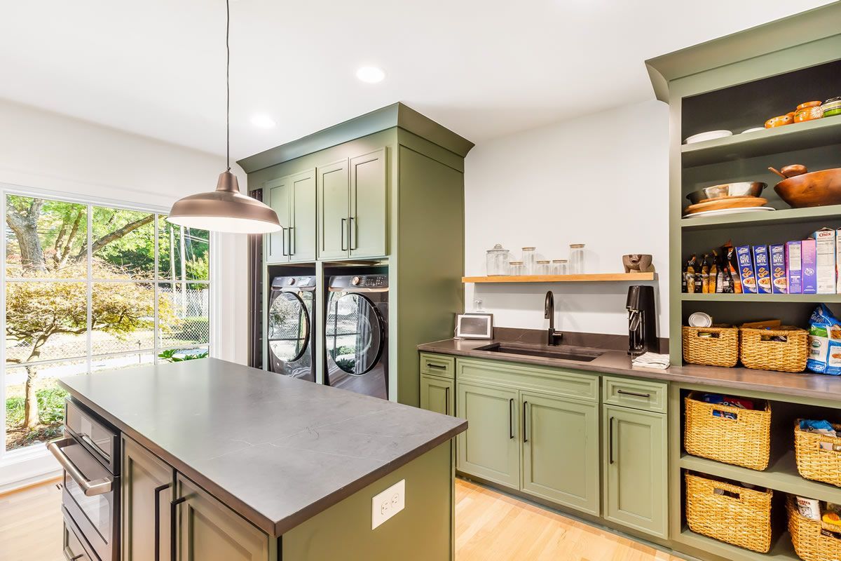 Kitchen with olive-green cabinets, a laundry area, and a large island. Sunlight streams through a window.