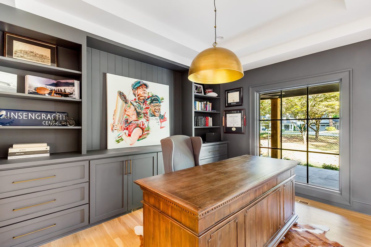 Home office with gray built-in shelving, wooden desk, gold pendant light, and colorful artwork.