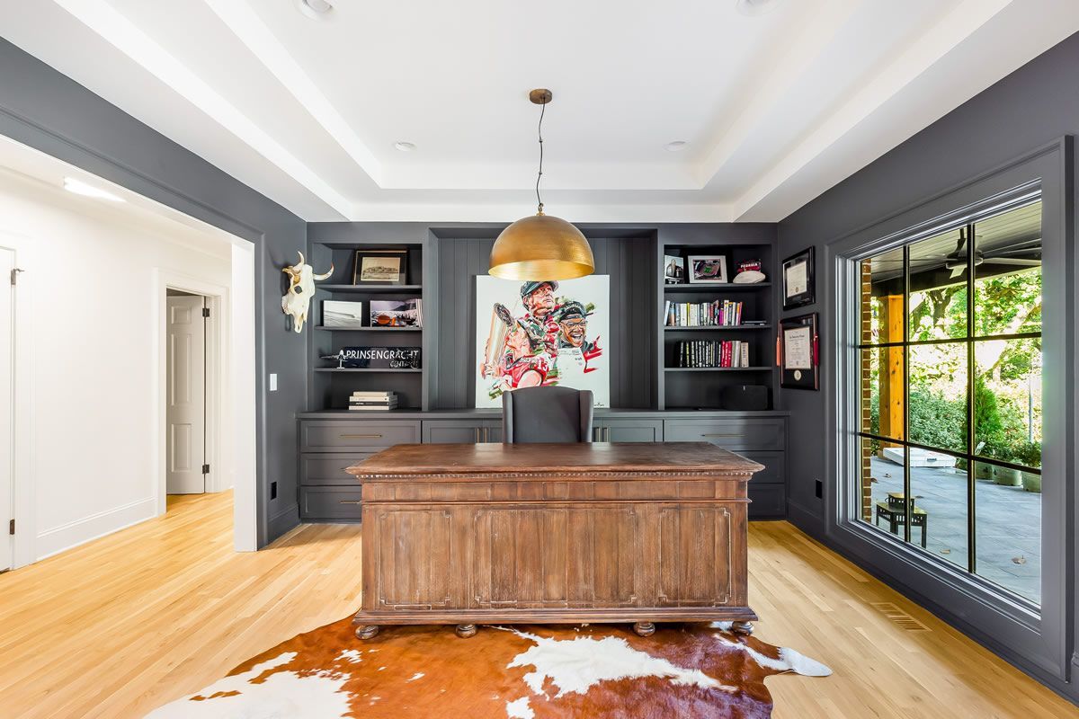 Office with dark gray walls, built-in shelving, wooden desk, cowhide rug, and large window overlooking a yard.
