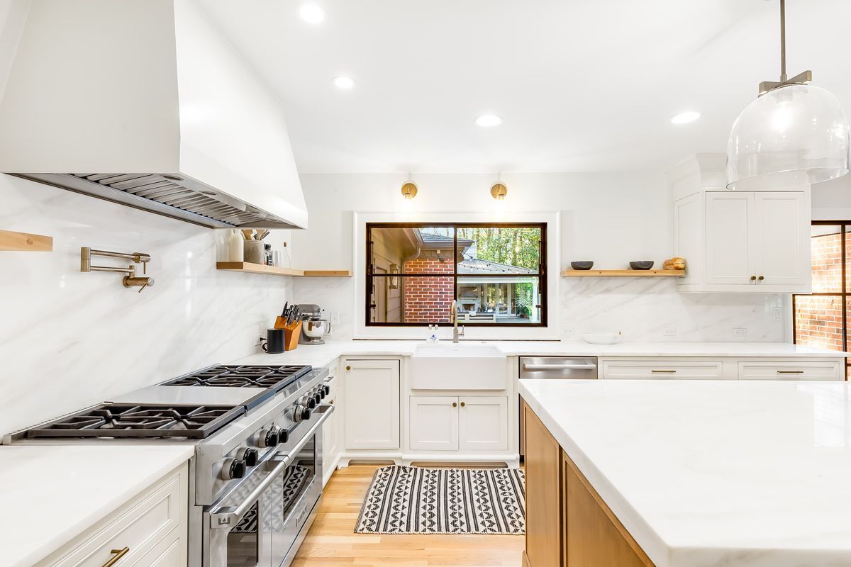 Bright, white kitchen with marble backsplash, island, stove, and sink. Wood accents and window with outdoor view.