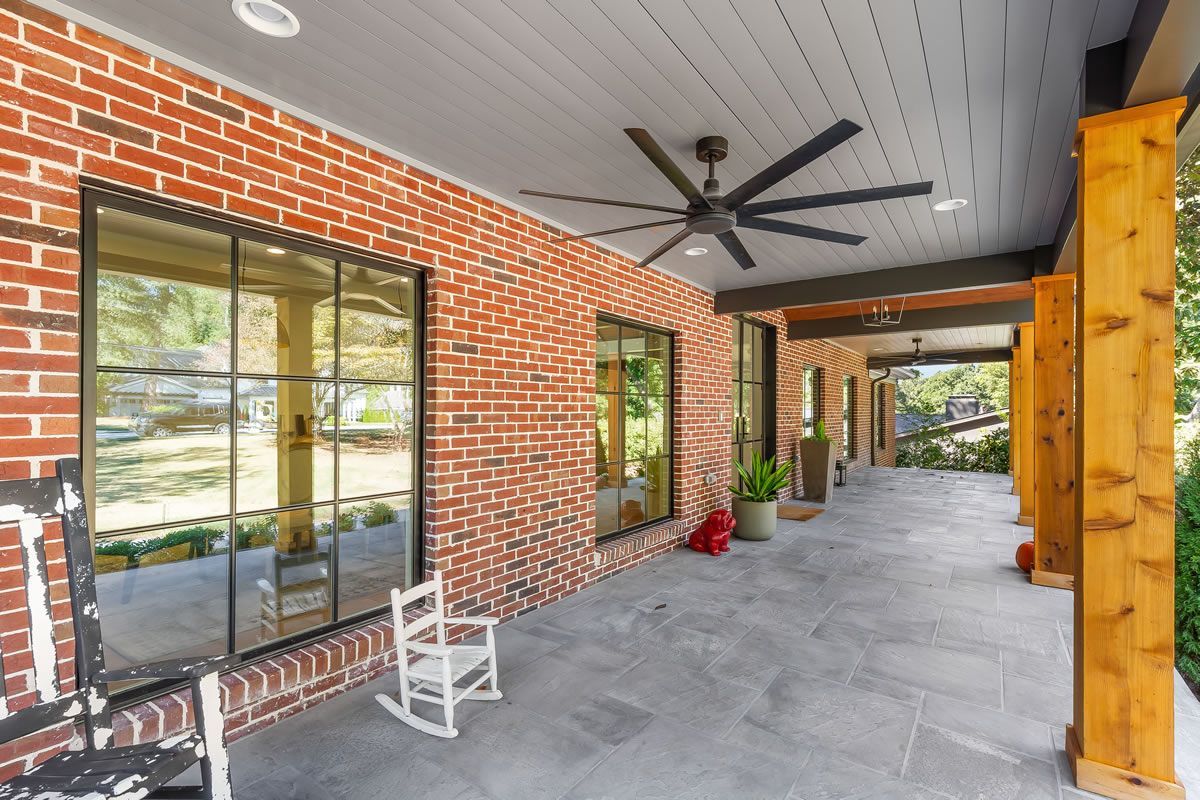 Covered porch with red brick wall, large windows, ceiling fan, and rocking chair.