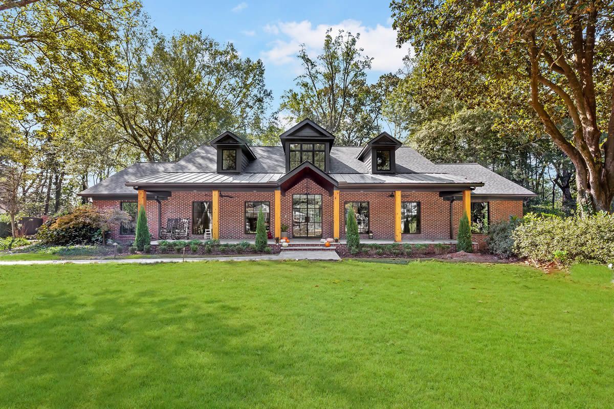 Brick house with a green lawn, wood columns, and dormer windows, surrounded by trees under a blue sky.