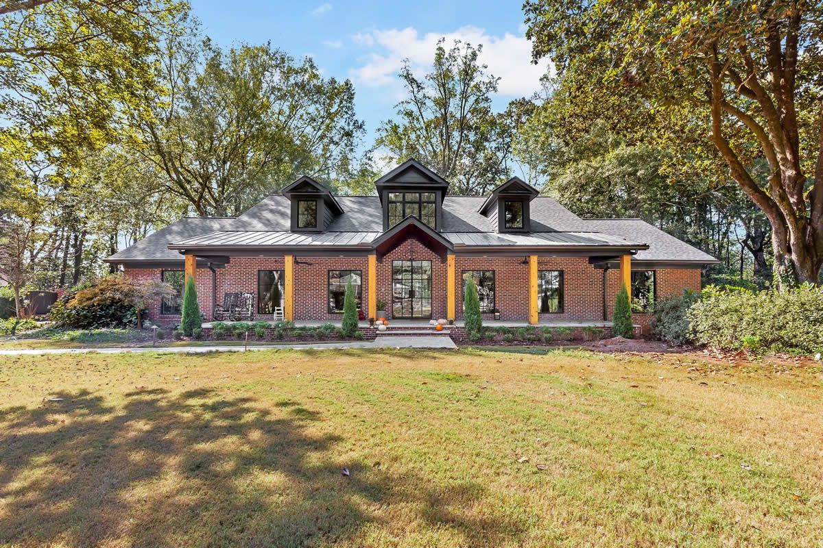 Brick ranch home with wooden columns and a metal roof, framed by trees and a green lawn.