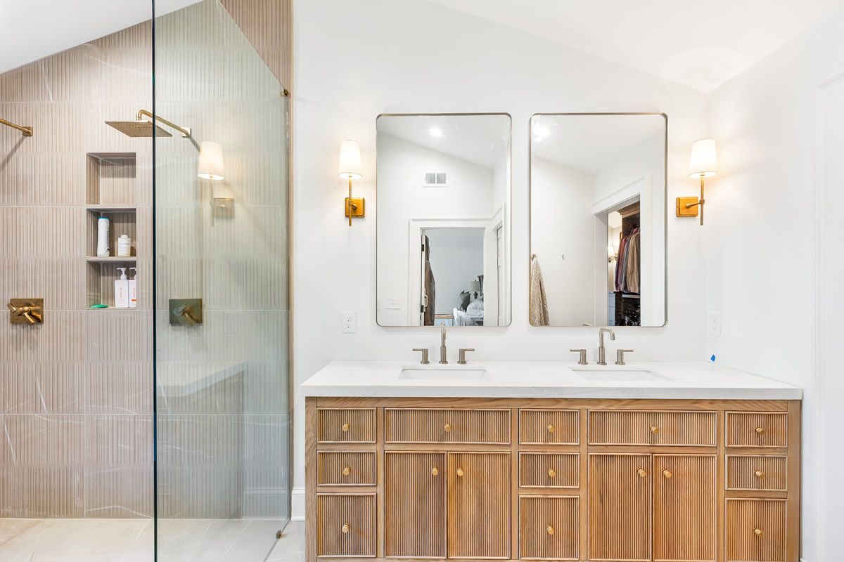 Bathroom with a light wood vanity, two mirrors, and a glass-enclosed shower with tile and gold fixtures.