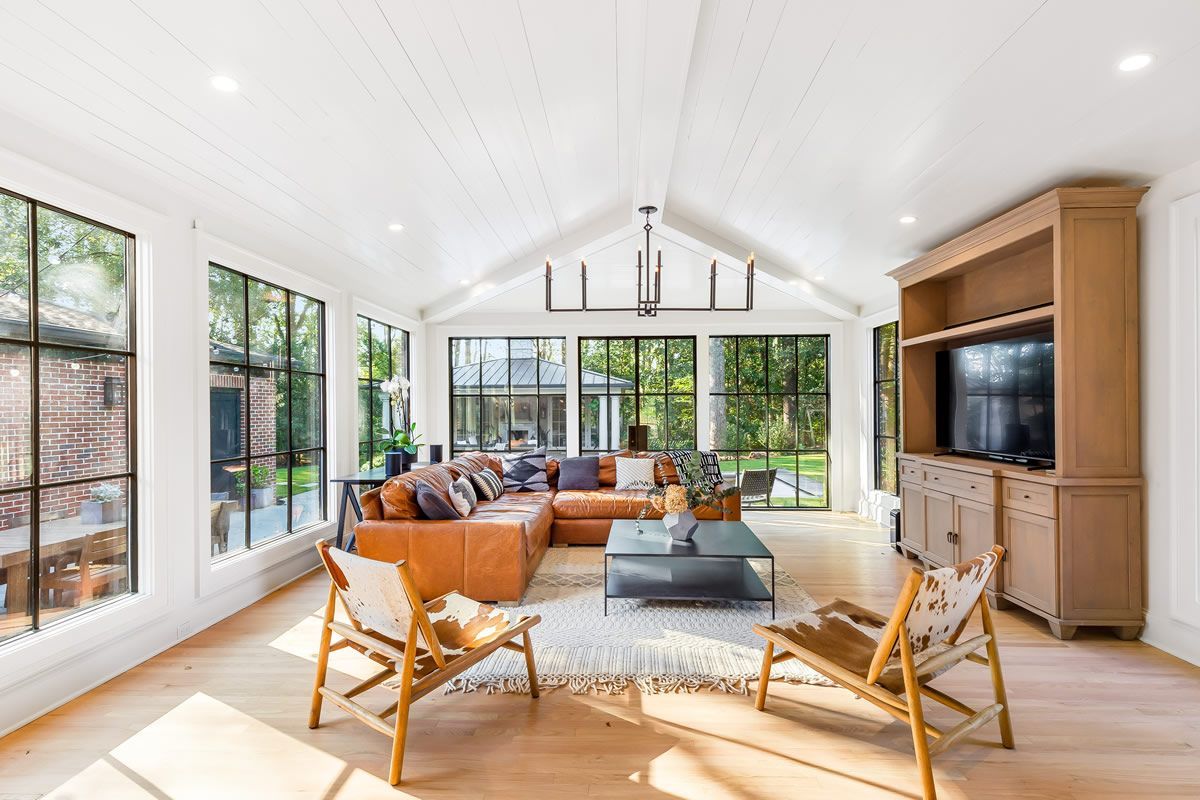 Bright, modern living room with leather sectional, floor-to-ceiling windows, and wooden chairs, flooded with sunlight.