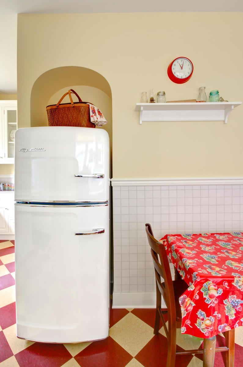 White vintage refrigerator in a kitchen with a red and white checkered floor and red floral tablecloth.