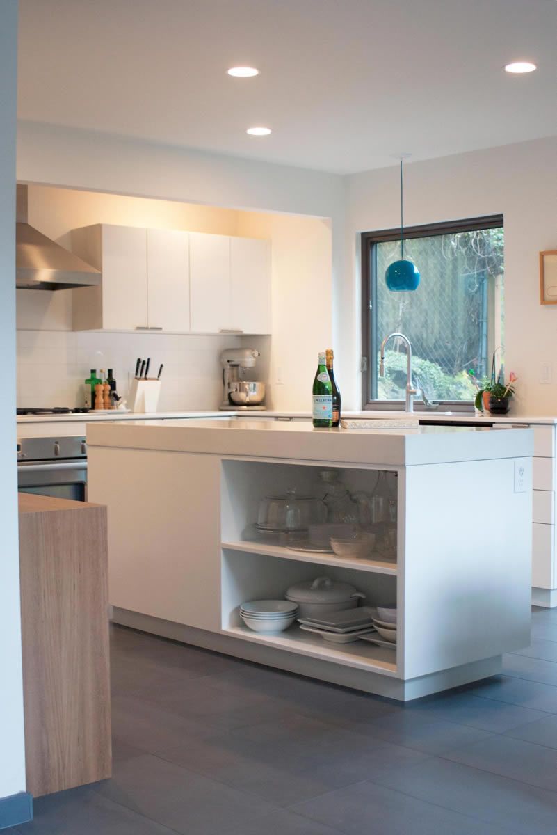 Modern white kitchen with island, cabinets, and window overlooking a garden.