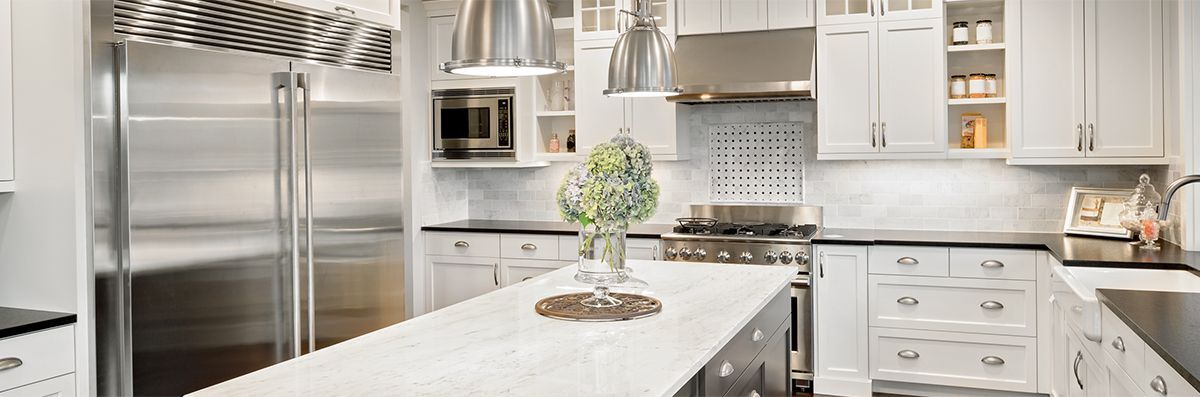 Modern white kitchen with stainless steel appliances, marble countertops, and a central island with a plant.