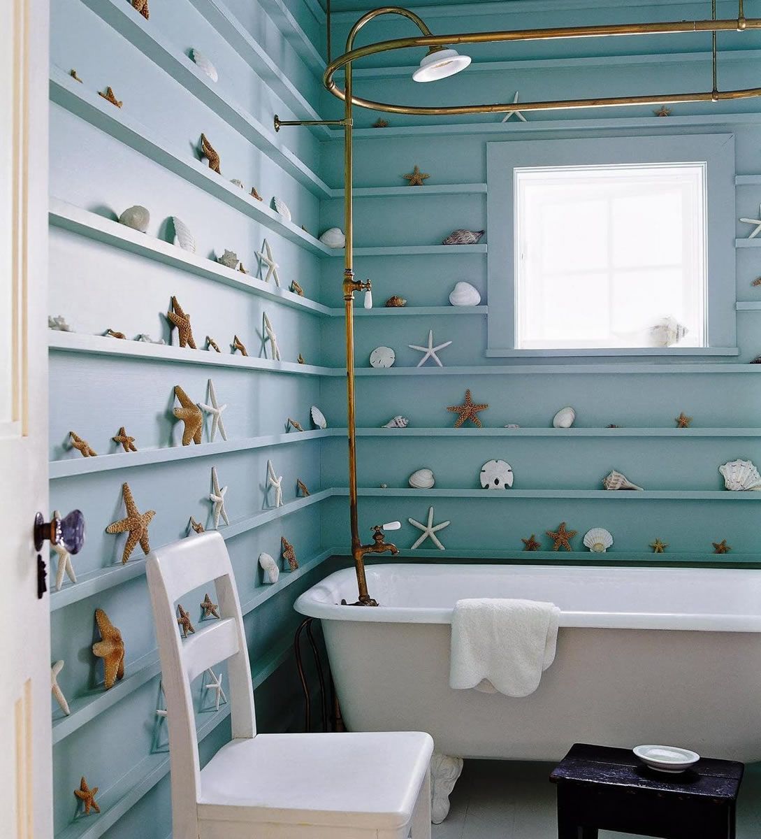 Blue bathroom with shelves of seashells, white chair, clawfoot tub, and brass shower fixtures.
