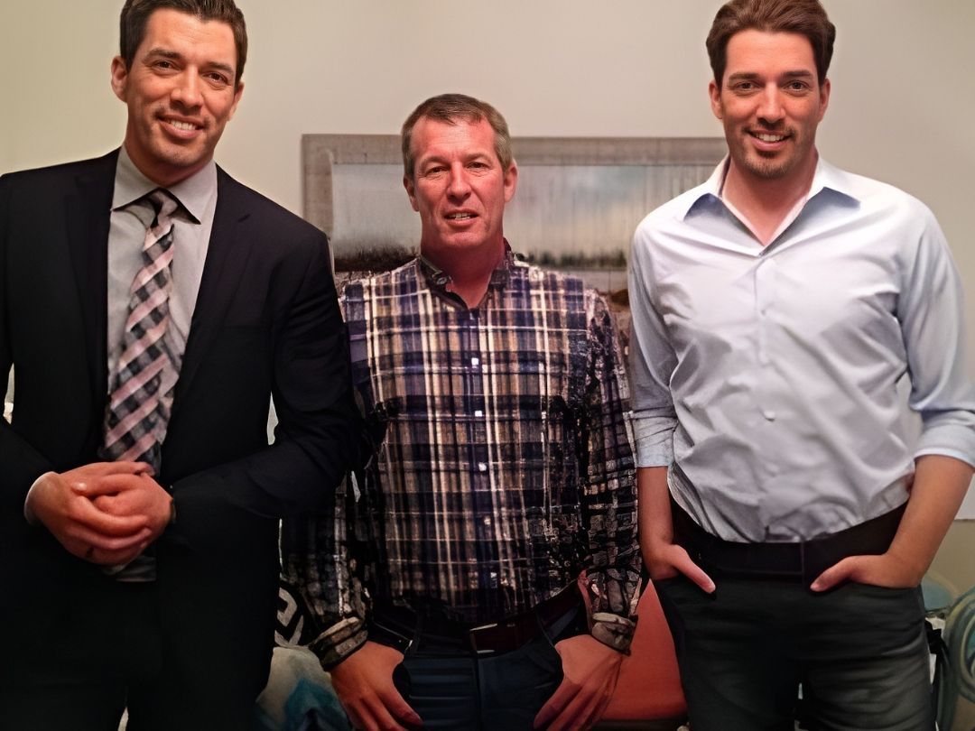Three men smiling, standing close together indoors. Two on the sides wear suits, the middle man wears a plaid shirt.