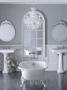 Elegant white bathroom with gray walls, clawfoot tub, pedestal sinks, ornate mirrors, and a chandelier.