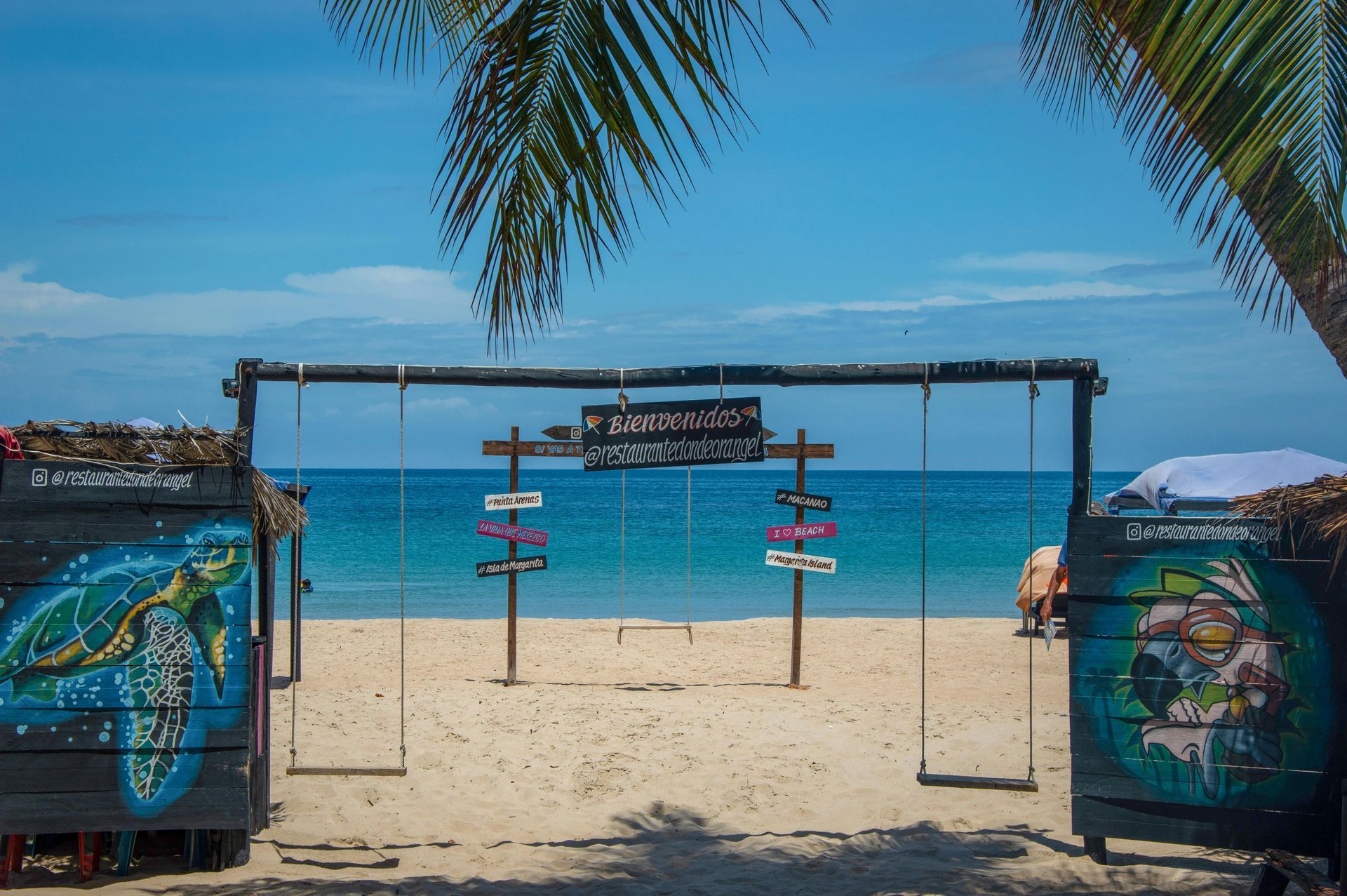 Entrada a la playa con coloridos letreros y vista al océano bajo un dosel de palmeras.