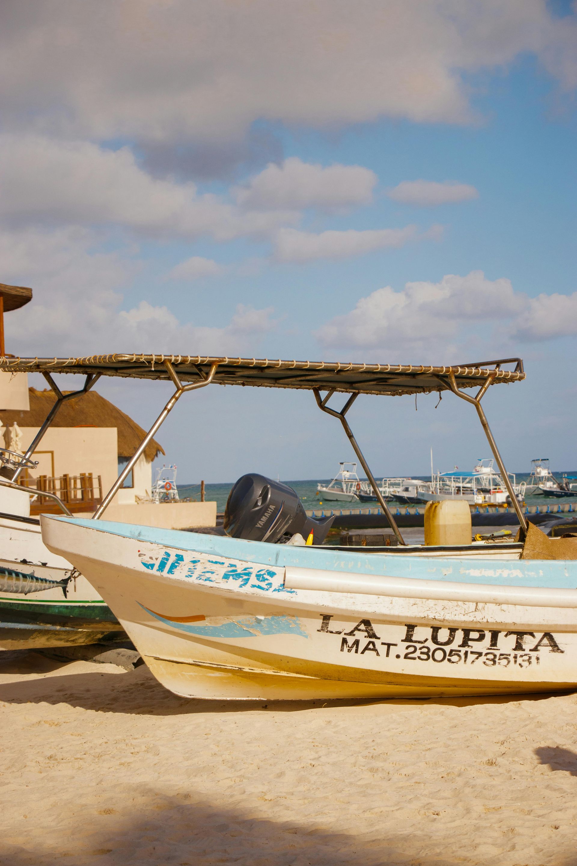 Una pequeña barca blanca en una playa de arena bajo un toldo, con cielo azul y nubes al fondo.