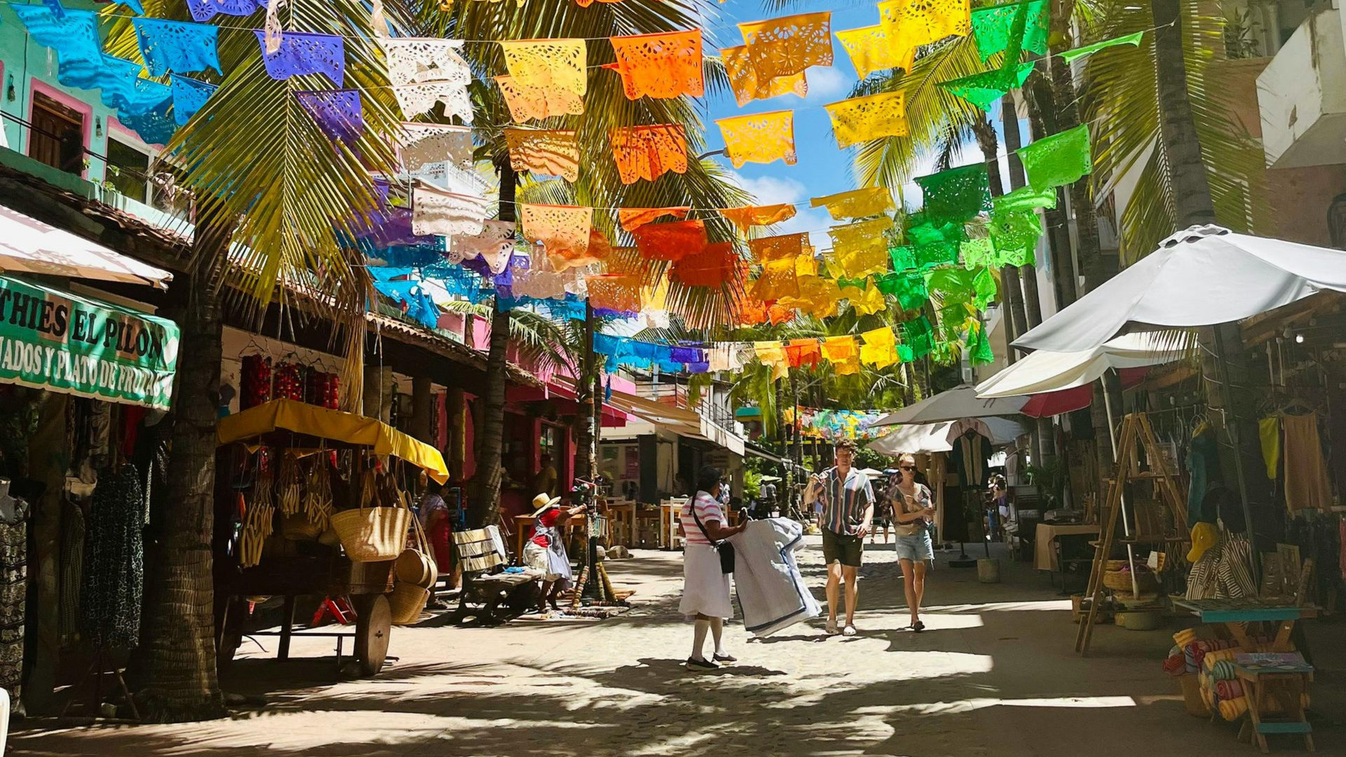 Banderines de colores ondean sobre una calle soleada del mercado, con tiendas, sombrillas y peatones.