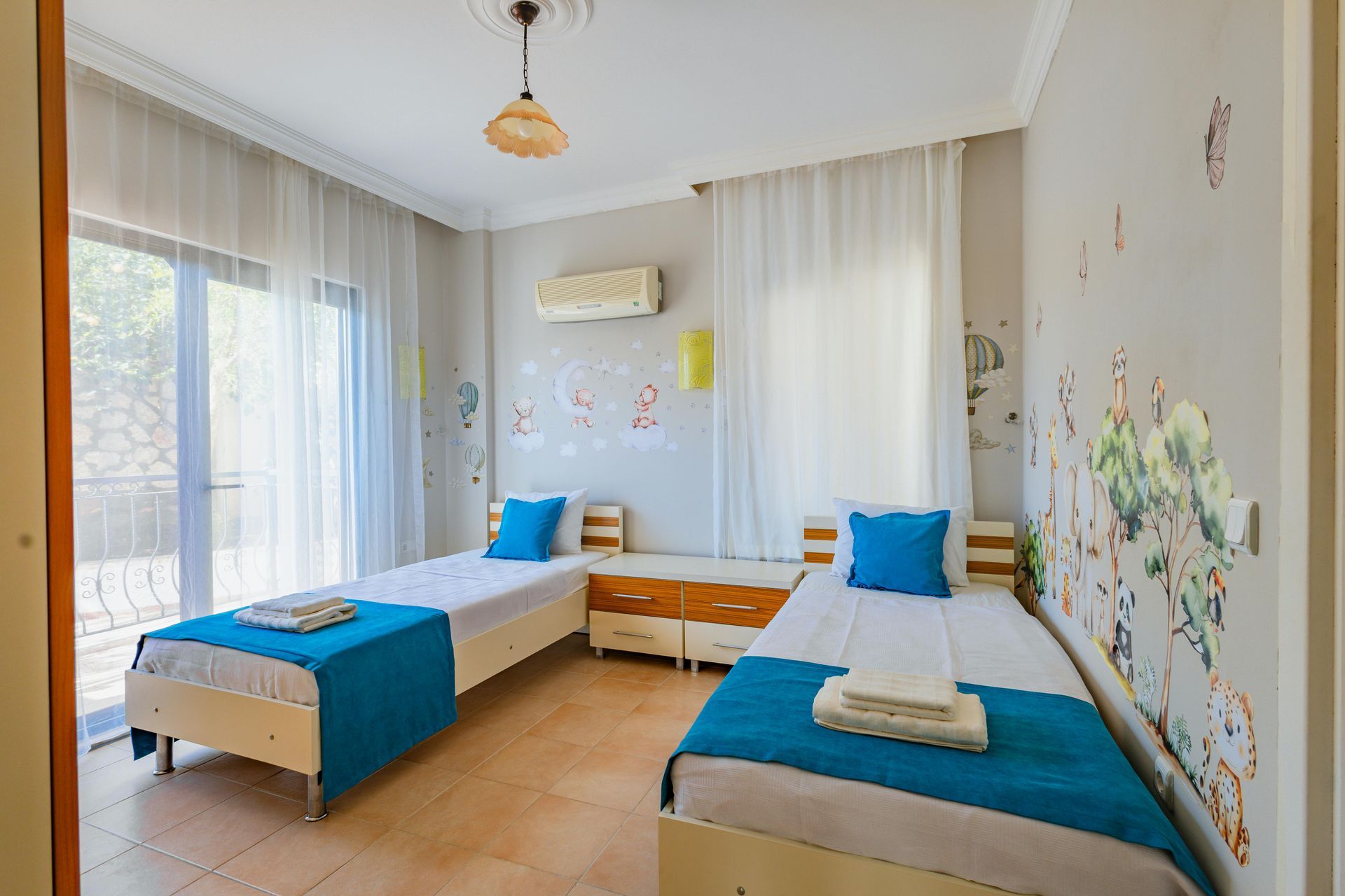 Habitación luminosa con dos camas individuales, colchas azules, cortinas blancas y vinilos decorativos florales en la pared.