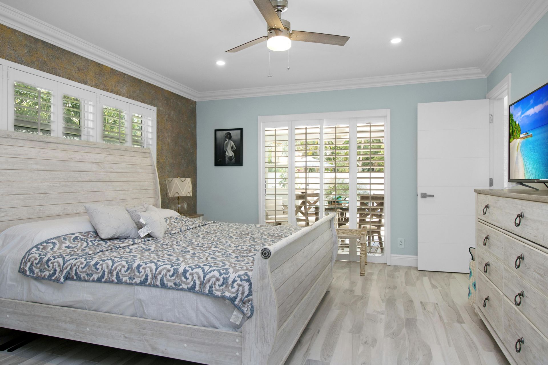 Habitación luminosa con cama gris, cómoda, ventilador de techo y puertas de cristal que dan a un patio.