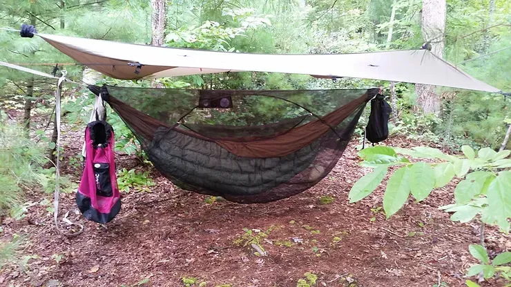 A hammock with a tarp and a backpack hanging from it in the woods.