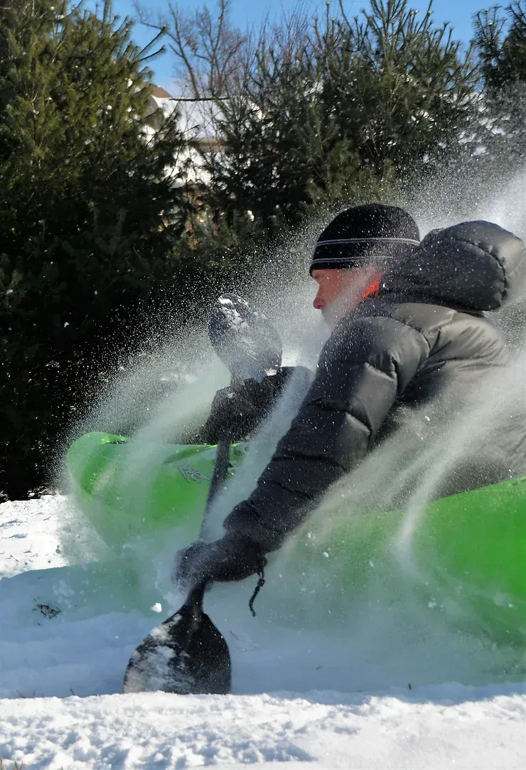 A man is riding a green tube through the snow while holding a shovel.