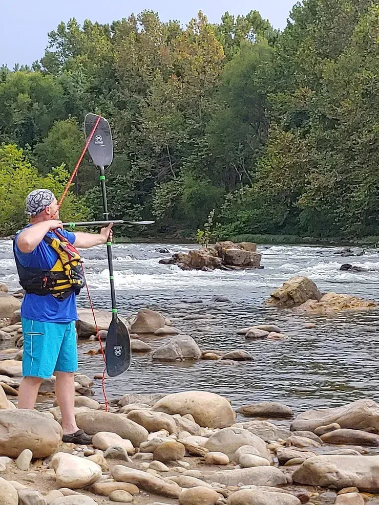 A man is standing on a rocky shore of a river holding a paddle.