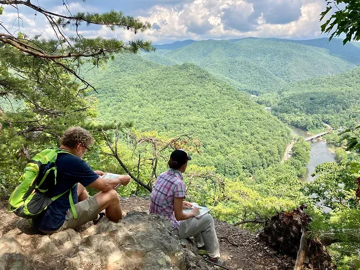 A man and a woman are sitting on a rock overlooking a river and mountains.