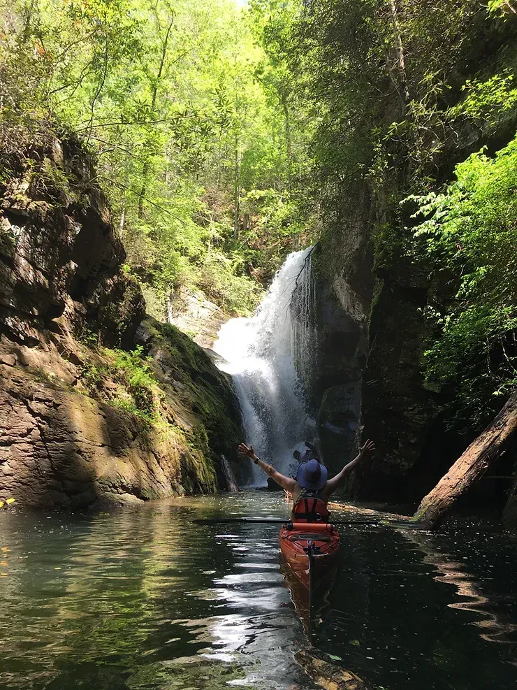 A man in a kayak is standing in front of a waterfall.