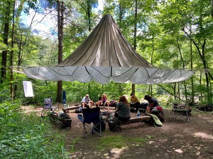 A group of people are sitting around a picnic table under a tent in the woods.