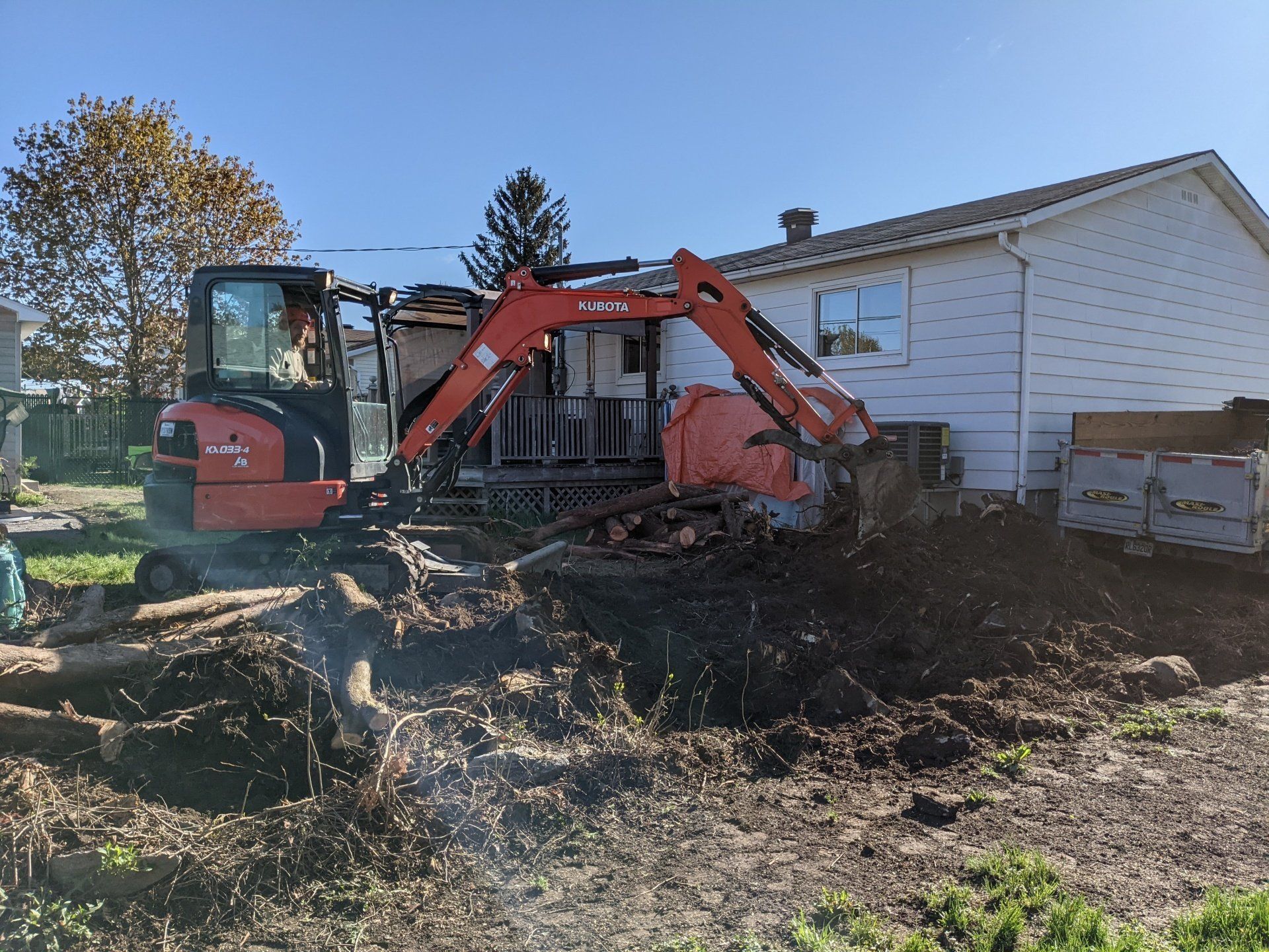 Une excavatrice Kubota orange démolissant une structure en bois à côté d'une maison blanche par une journée ensoleillée.