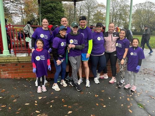 Group of people wearing purple shirts with a daisy logo, smiling outdoors in a park, possibly for a charity event.