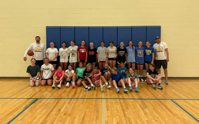 Youth volleyball team posing in a gym, with players in jerseys and coaches standing in front of blue wall pads.