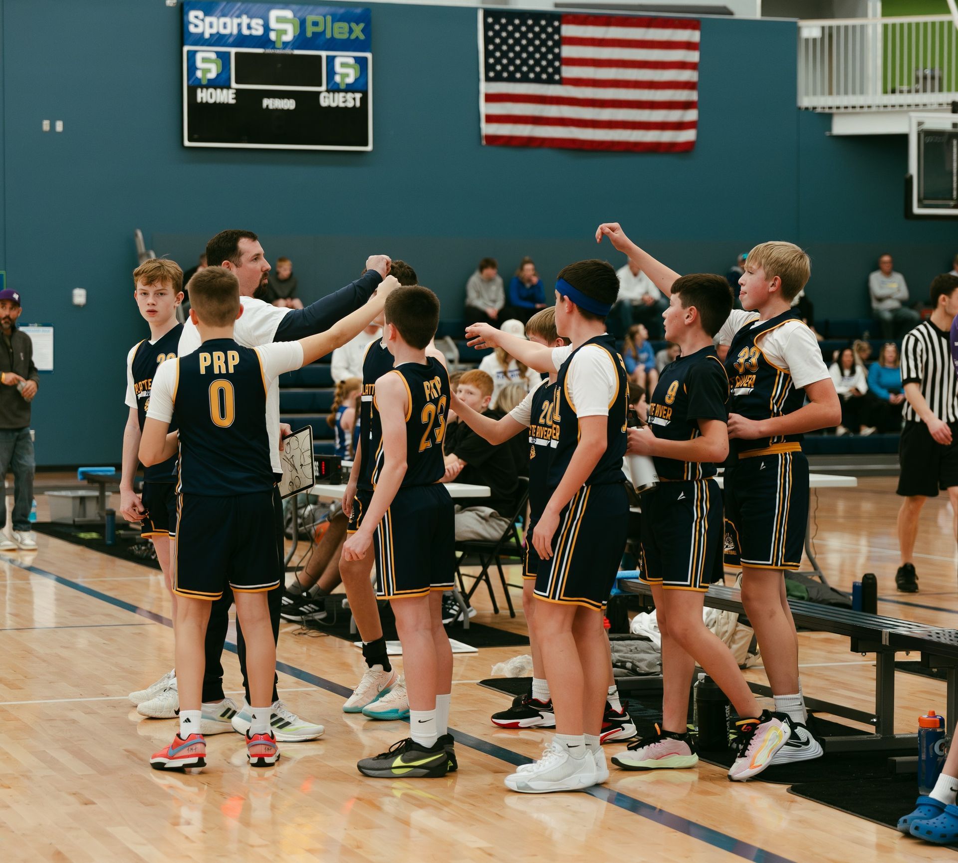 Youth basketball team huddled on court near bench, wearing black and gold uniforms with an American flag above.